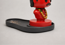 A red and black stylized bust figurine with a horn-like hairpiece, shown on a black tray with incense sticks and rising smoke against a light gray studio background.